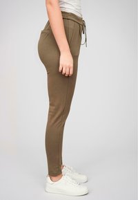 Olivenfargede slim-fit joggebukser laget av elastisk materiale, med snor i livet, sidelommer og splittede sømmer, kombinert med hvite sneakers.