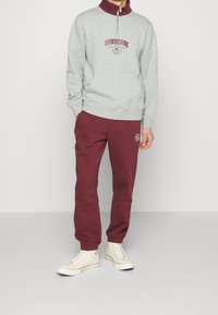 Grauer Fleece-Sweatshirt mit Viertelreißverschluss und burgunderfarbenen Akzenten, kombiniert mit burgunderfarbenen Jogginghosen mit einem Logo. Weiße High-Top-Sneaker.
