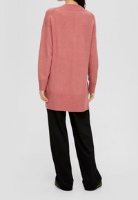 Pull long en maille rose avec ourlet et poignets côtelés, offrant une coupe décontractée et des fentes sur les côtés, associé à un pantalon noir à jambes larges.