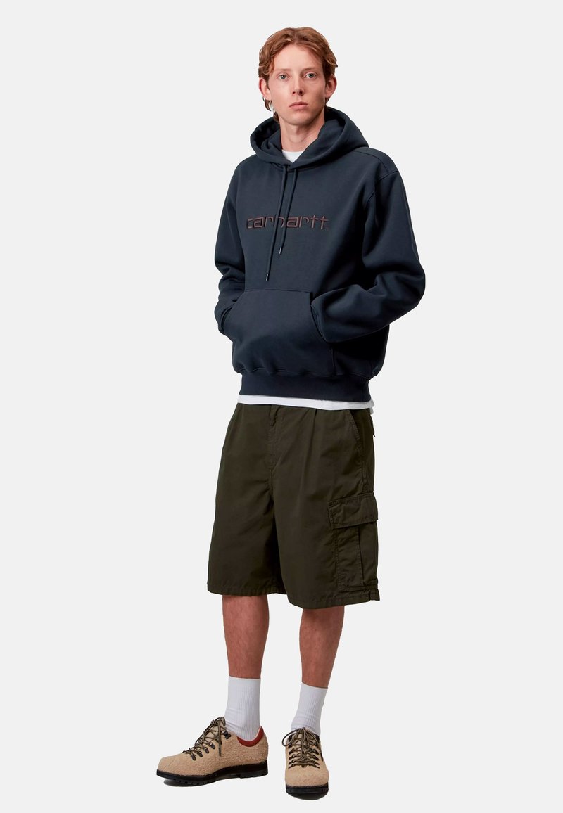 Felpa con cappuccio blu navy presenta una tasca frontale e un logo; abbinata a pantaloni cargo color oliva e scarpe beige con suole e lacci scuri.