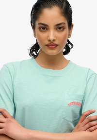 Lys turkise t-shirt med korte ærmer, der har en tekstureret overflade og en rød broderet "SUPERDRY" på venstre brystlomme.