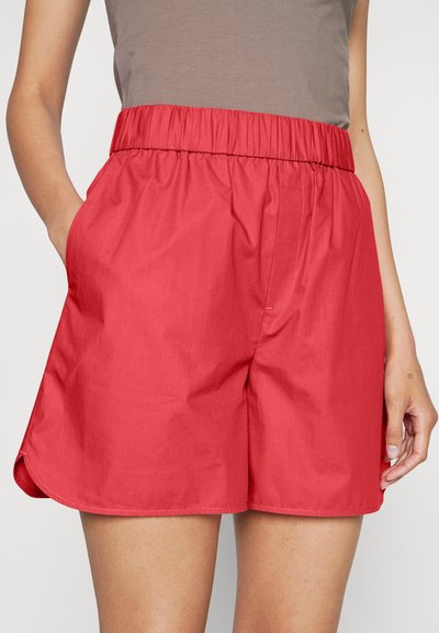 En kvinne iført høyt liv rød shorts med elastisk midjebånd og sidelommer, parret med en grå topp som er stoppet inn.