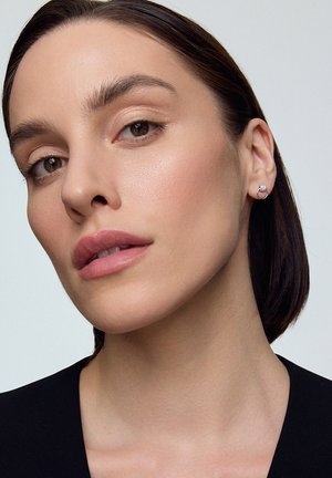 Srebrna kolczyk w kształcie obręczy z okrągłym akcentem gemu, noszony na uchu modelki. Gładka skóra, neutralny makijaż i ciemne, proste włosy. Czarna bluzka.