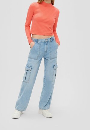 Vrouw die een koraalroze cropped top met lange mouwen draagt, lichtblauwe wijde cargo-spijkerbroek en witte sneakers, staand met handen in de zakken.