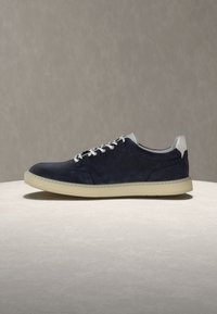 Baskets en daim bleu marine avec lacets blancs, languette de talon blanche, et semelle en caoutchouc beige sur une surface claire contre un fond gris.