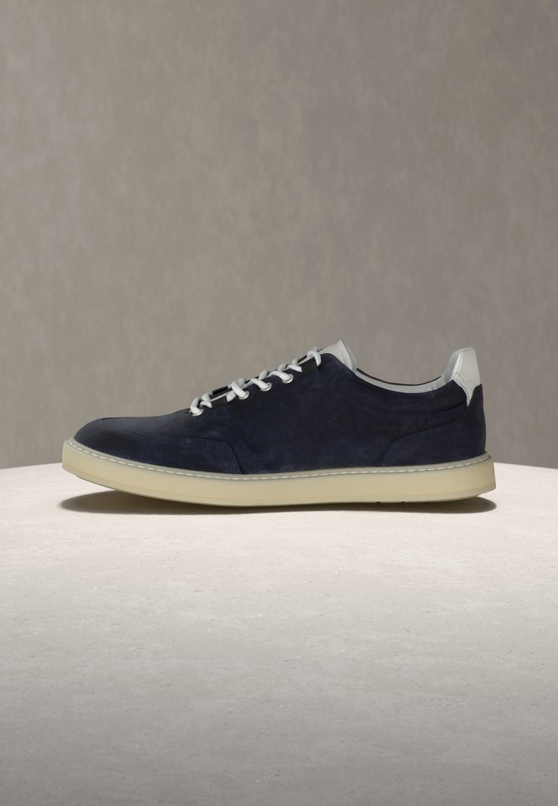 Baskets en daim bleu marine avec lacets blancs, languette de talon blanche, et semelle en caoutchouc beige sur une surface claire contre un fond gris.