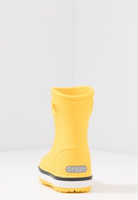 Botte en caoutchouc jaune avec un tige haute, intérieur texturé et un accent de logo gris sur le talon. Présente une semelle rayée blanche et marine.