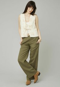 Gilet sans manches blanc cassé en tissu texturé avec des motifs carrés, noué à l'avant ; associé à un pantalon ample olive et des bottines brunes.