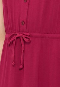 Μπορντό φόρεμα με κουμπιά στο μπροστινό μέρος και ζώνη από ύφασμα που δένει στη μέση.