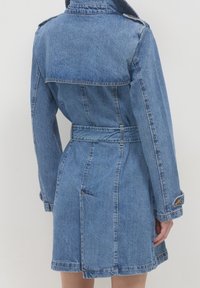 Denim jacket in light blue with a belted waist, fold-over collar, and shoulder epaulets. Features prominent seams and two side pockets.