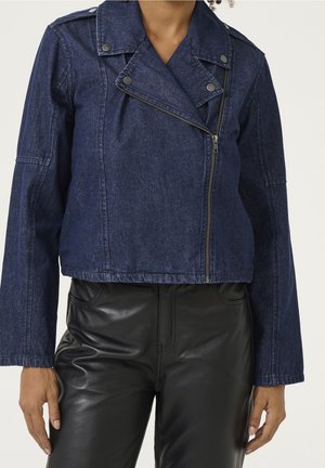 Femme portant une veste courte en jean bleu foncé avec une fermeture éclair diagonale et un pantalon en simili cuir noir sur fond uni.
