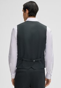 Dunkelgrauer Weste mit glatter, strukturierter Oberfläche. Verfügt über ein V-Rücken-Design und einen verstellbaren Riemen. Wird über einem weißen Langarmshirt getragen.