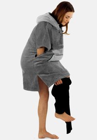 Grau-hooded Poncho mit Handtuchtextur. Mit seitlichen Schlitzen und einer kontrastierenden dunklen Tasche. Die Person scheint schwarze Leggings hochzuziehen.