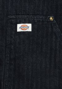 Τσέπη από τζιν ένδυμα με ορατή λευκή ετικέτα της μάρκας Dickies και χρυσό καρφί στη πάνω δεξιά γωνία της τσέπης.