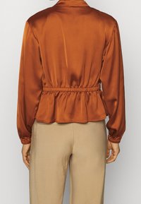 Rustrød satinbluse med peplum-design, samlet talje og lange ærmer. Den glatte tekstur og lette glans fremhæver formen.