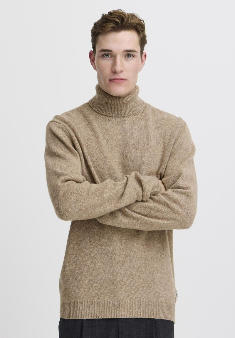 Maglione dolcevita beige in tessuto morbido a maglia, con maniche lunghe e collo a coste, indossato da una modella con le braccia incrociate.