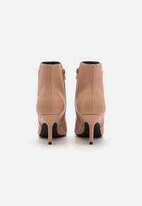 Botas de tobillo de cuero beige con un diseño elegante, cremallera lateral, punta afilada y un fino tacón negro alto. Textura suave, apariencia minimalista.