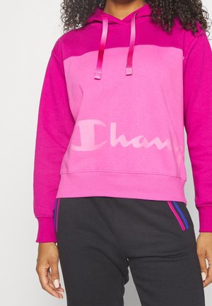 Frau trägt einen rosa- und magentafarbenen Hoodie mit „Champion“-Logo auf der Vorderseite und schwarze Hose mit pink-blau gestreiften Taschen.