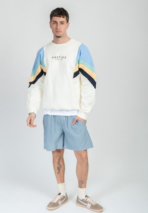 Young man wearing white sweatshirt with blue, green, yellow, and black stripes, blue shorts, beige sneakers, standing against plain gray background.