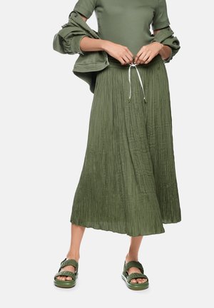 Frau trägt olivgrünen, plissierten Midi-Rock mit Kordelzug, passende Sandalen und darüber eine aufgekrempelte Jacke über einem kurzärmeligen Oberteil, steht vor weißem Hintergrund.