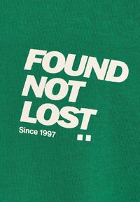 Texto blanco "ENCONTRADO, NO PERDIDO Desde 1997" impreso en diagonal sobre fondo de tela de punto verde.