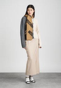 Sokszínű pulóver szürke és krémszínű kötött mintákkal, bézs, földig érő szoknya és barna sál fekete logóval. Fekete szandál fehér zoknival.