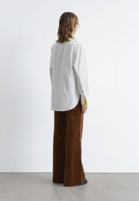 Loose-fitting white shirt with a rounded hem, paired with brown wide-leg corduroy pants. Floor-length, smooth texture and simple design.