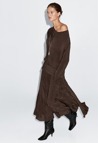 Brązowa bluzka na jedno ramię z rozwianą spódnicą, wykonana z miękkiego materiału. W zestawie eleganckie czarne botki do kolan. Prosta srebrna naszyjnik z wisiorkiem.