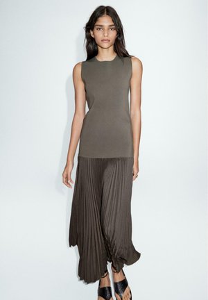 Olivengrünes ärmelloses Oberteil kombiniert mit dunklen, plissierten weit geschnittenen Hosen. Das Outfit hat eine glatte Textur und eine lässige Passform. Schwarze flache Sandalen runden den Look ab.