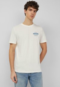 Młody mężczyzna stoi oparty o szarą ścianę, ubrany w białą koszulkę z krótkim rękawem z małym niebieskim napisem na piersi oraz jasnoniebieskie dżinsy.