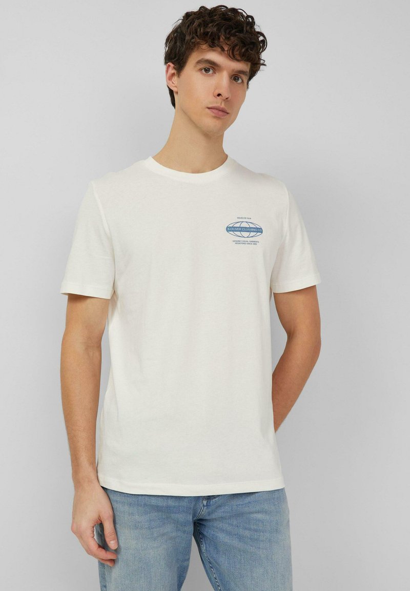 Młody mężczyzna stoi oparty o szarą ścianę, ubrany w białą koszulkę z krótkim rękawem z małym niebieskim napisem na piersi oraz jasnoniebieskie dżinsy.