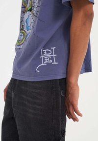 Lilla bomulds t-shirt med et grafisk design på fronten og hvid tekst på sidekant. Sammen med mørke denim jeans.