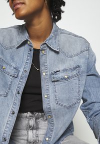 Jasnoniebieska koszula denimowa z guzikami z przodu, dwoma kieszeniami na piersi i starannym przeszyciem. Nosi się ją na czarnym topie, w połączeniu z szarymi spodniami.