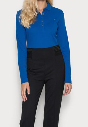 Kobieta ubrana w niebieską koszulkę polo z długim rękawem oraz czarne spodnie z wysokim stanem na jednolitym jasnym tle.