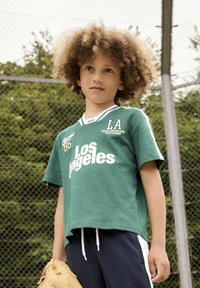 Grøn kortærmet t-shirt med "Los Angeles" og "LA" tryk, parret med navy bukser med hvide sidelæg; holder en beige baseballhandske.