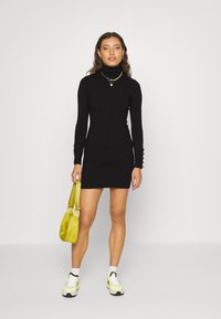 Robe pull noire côtelée avec un col haut et des manches longues ornées de boutons. Associée à un sac vert clair et des baskets.