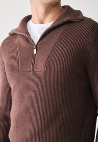 Pull en tricot côtelé marron avec un col montant et une fermeture éclair à mi-longueur. Le tissu texturé présente un motif en relief sur la poitrine.