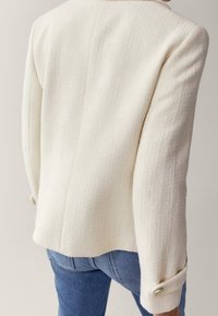 Blazer bianco testurizzato con una forma aderente, maniche lunghe e polsini con bottoni, abbinato a jeans blu. La vista posteriore evidenzia il materiale e il design.