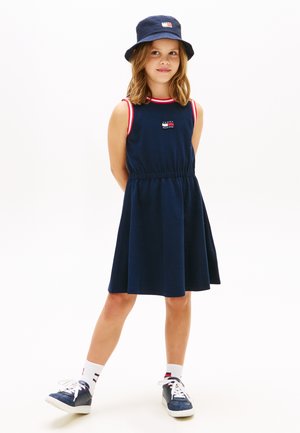 Fiatal lány tengerészkék ruhában, piros és fehér szegéllyel, tengerészkék kalapban, fehér zokniban és tengerészkék tornacipőben, kezét a háta mögött tartva áll.