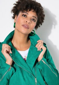 Giacca in suede verde con colletto, zip frontale e tasche laterali. Il modello tiene il colletto; il tessuto testurizzato è visibile.