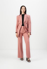 Růžový na míru šitý blazer a široké kalhoty z matného materiálu, v kombinaci s černým přiléhavým topem a černými sandály na podpatku. Detail brože na blazeru.