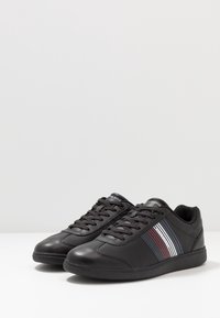 Zwarte sneakers van glad leer, met een ronde neus, gestructureerde zool en drie horizontale strepen in rood, wit en blauw aan de zijkant.