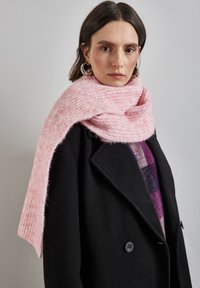 Écharpe en laine texturée rose drapée sur un manteau noir, présentant un design à double boutonnage et un bouton. L'écharpe a une apparence douce et duveteuse.