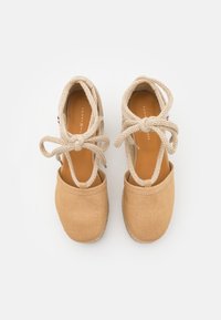 Tan canvas espadrilles met beige touwtjes, open zijkanten en een textuur van jute zolen. Aan de binnenkant bevindt zich een bruine binnenzool met branding.