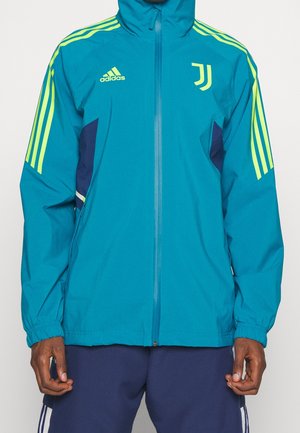 Homme portant une veste zippée turquoise Adidas avec des bandes jaune néon sur les manches et le logo de la Juventus sur la poitrine.