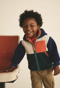 Fleece bunda s dizajnom farebných blokov v teal, bielej, námorníckej modrej a oranžovej; má zips a kapucňu, nosená cez červené tričko a khaki nohavice.