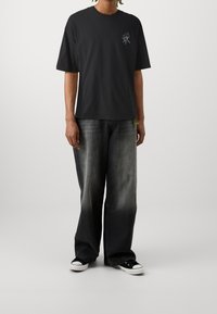 Svart t-shirt med kort ärm och lotusblomma broderad på bröstet, tillsammans med löst sittande, urtvättade svarta jeans och svarta sneakers.