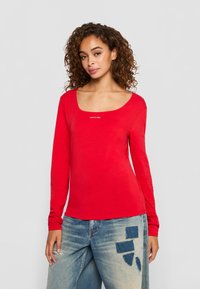 Vrouw met krullend haar, gekleed in een rode long-sleeve top en een blauwe jeans met denim patches, staand tegen een effen lichte achtergrond.
