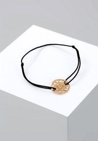 Schwarzes Kordelarmband mit einem kreisförmigen, goldfarbenen Metallanhänger mit geometrischem Muster und zentralem Blumendesign.