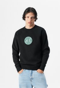Črna trenerka iz bombaža s krožnim izrezom, ki ima na prsih zeleno okroglo logo in rebraste manšete, v kombinaciji z svetlo modrimi jeans hlačami.